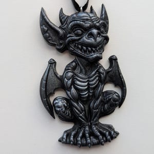 May include: A dark gray gargoyle pendant with a detailed, textured design. The pendant features a winged creature with horns, large ears, and a menacing grin. The gargoyle is suspended from a black cord, suitable for wearing as a necklace.