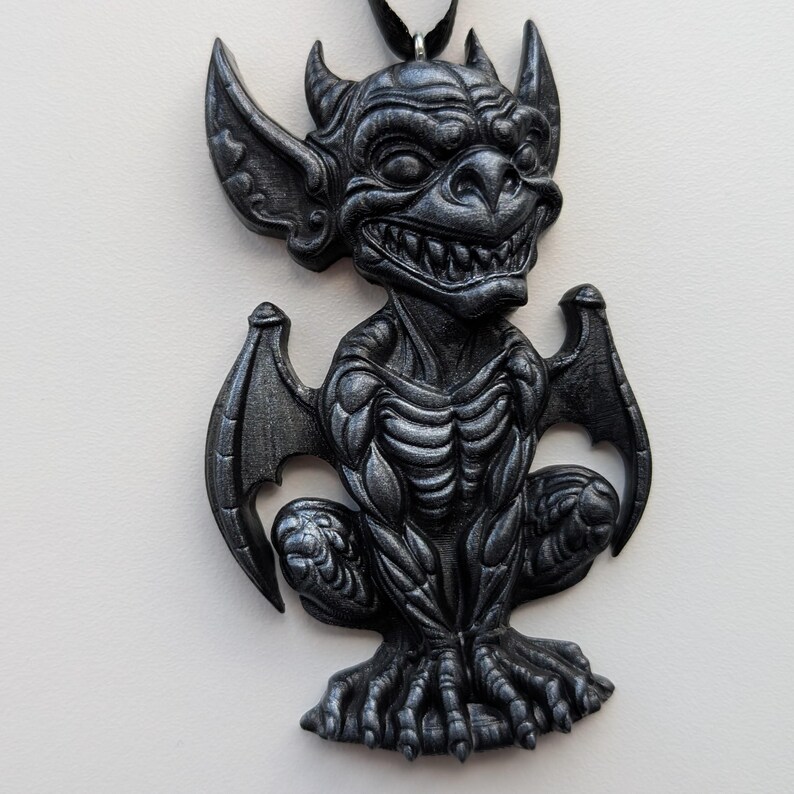 May include: A dark gray gargoyle pendant with a detailed, textured design. The pendant features a winged creature with horns, large ears, and a menacing grin. The gargoyle is suspended from a black cord, suitable for wearing as a necklace.