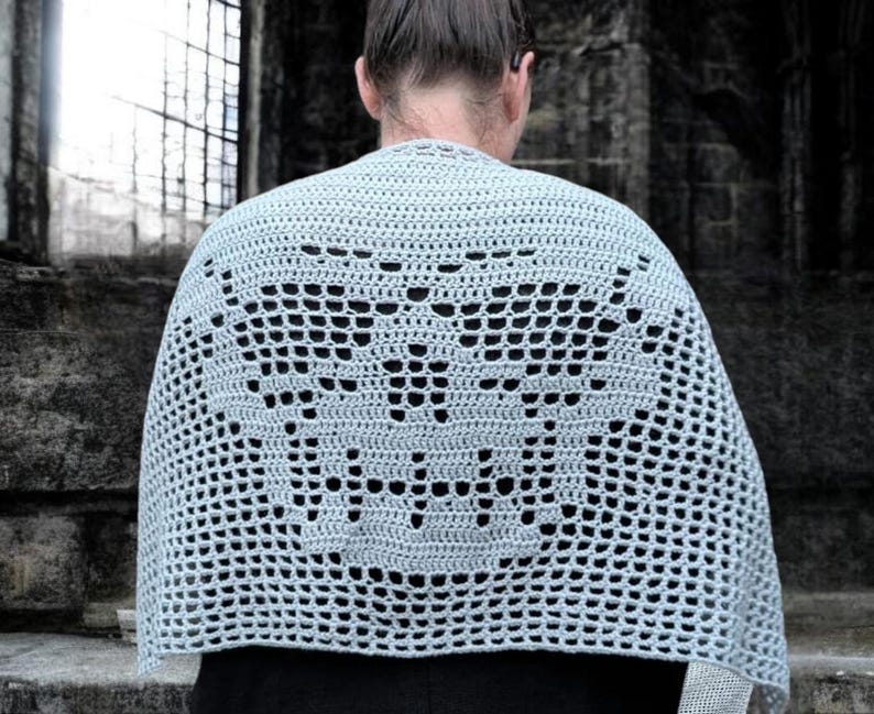 May include: A light gray crocheted shawl with a decorative openwork pattern. The shawl is draped over a person's shoulders, with the back of the person facing the camera. The shawl's design is visible, and the background is a stone structure.