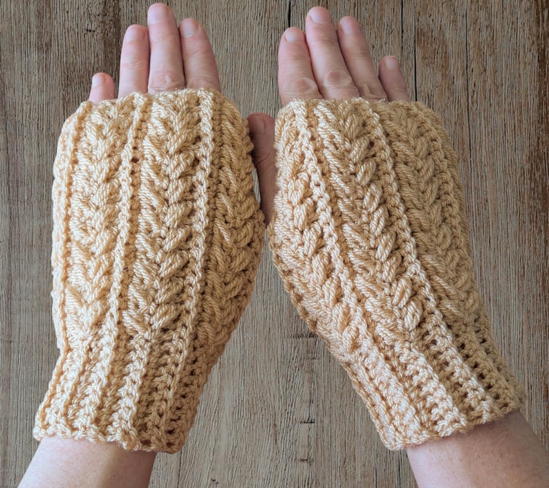 May include: A pair of beige fingerless gloves with a textured, woven pattern. The gloves are made of a soft yarn and have a ribbed cuff. The design features a repeating leaf-like motif.