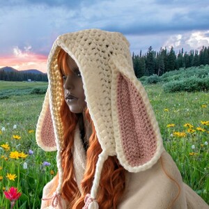 May include: A white crocheted hooded hat with pink bunny ears. The hat is worn by a person with long red hair. The person is sitting in a field of flowers.