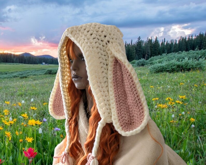 May include: A white crocheted hooded hat with pink bunny ears. The hat is worn by a person with long red hair. The person is sitting in a field of flowers.