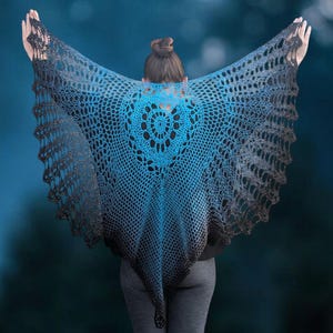 May include: A handmade, open-weave shawl in shades of blue and gray, with a heart-shaped design at the center. The shawl is draped over the shoulders, creating a wing-like effect. The background is a blurred, dark blue with hints of light.