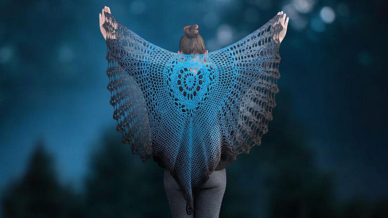May include: A handmade, open-weave shawl in shades of blue and gray, with a heart-shaped design at the center. The shawl is draped over the shoulders, creating a wing-like effect. The background is a blurred, dark blue with hints of light.