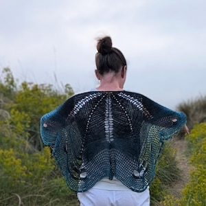 Puede incluir: Un chal hecho a mano, de tejido abierto, en tonos azules y negros, con efecto degradado. El chal se lleva sobre una camiseta blanca, con un dise&ntilde;o abierto &uacute;nico. Los bordes del chal son de color turquesa brillante y el estilo general es bohemio.