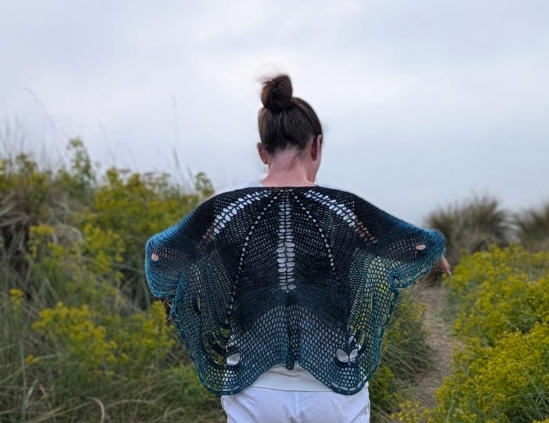 Puede incluir: Un chal hecho a mano, de tejido abierto, en tonos azules y negros, con efecto degradado. El chal se lleva sobre una camiseta blanca, con un dise&ntilde;o abierto &uacute;nico. Los bordes del chal son de color turquesa brillante y el estilo general es bohemio.