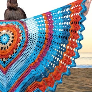 May include: A colorful, crocheted shawl with a circular design in shades of orange, tan, and blue. The shawl has a scalloped edge and is held up against a beach backdrop, showcasing its intricate openwork pattern.