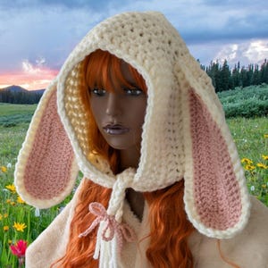 May include: A white crocheted bunny hood with pink ears. The hood has a long tie that can be worn around the neck. The hood is worn by a person with long red hair. The person is standing in a field of flowers.