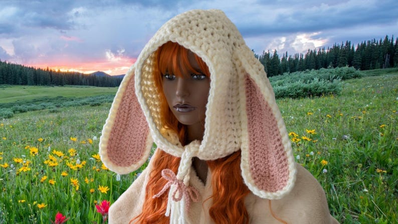 May include: A white crocheted bunny hood with pink ears. The hood has a long tie that can be worn around the neck. The hood is worn by a person with long red hair. The person is standing in a field of flowers.