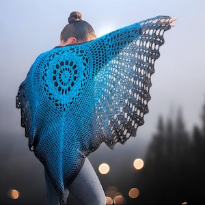 May include: A blue and black crochet shawl with a circular design on the back. The shawl is draped over the shoulders, with the edges flowing outward. The background is a blurred outdoor scene with trees.