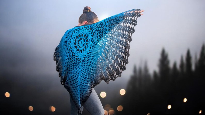 May include: A blue and black crochet shawl with a circular design on the back. The shawl is draped over the shoulders, with the edges flowing outward. The background is a blurred outdoor scene with trees.