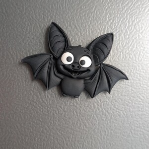 May include: A black bat-shaped decorative magnet with large, expressive eyes and a friendly smile. The bat has large ears and outstretched wings, giving it a cartoonish appearance. The magnet is likely made of a durable material.