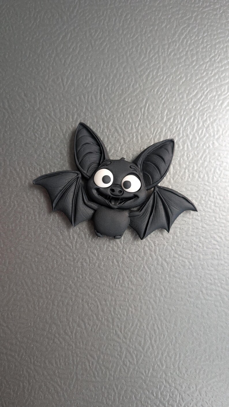 May include: A black bat-shaped decorative magnet with large, expressive eyes and a friendly smile. The bat has large ears and outstretched wings, giving it a cartoonish appearance. The magnet is likely made of a durable material.
