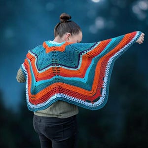 Puede incluir: Un chal de ganchillo hecho a mano con colores vibrantes, en tonos turquesa, naranja y blanco. El chal presenta un dise&ntilde;o geom&eacute;trico de estrella. La persona lleva una camisa verde de manga larga y vaqueros oscuros.