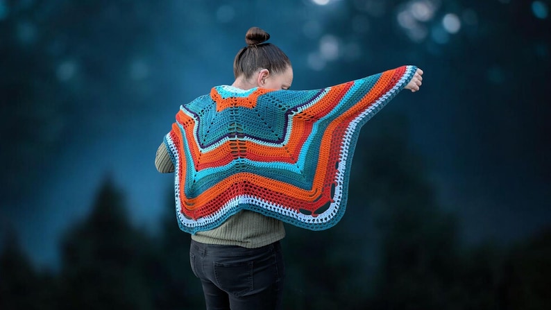 Puede incluir: Un chal de ganchillo hecho a mano con colores vibrantes, en tonos turquesa, naranja y blanco. El chal presenta un dise&ntilde;o geom&eacute;trico de estrella. La persona lleva una camisa verde de manga larga y vaqueros oscuros.