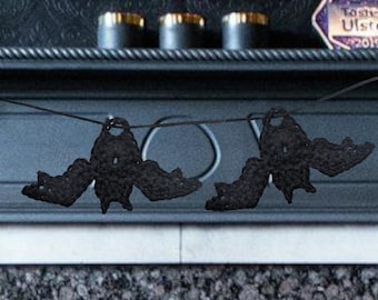 May include: Two black crochet bat decorations hanging on a string. The bats have outstretched wings and are suspended against a dark background. The bats are likely handmade and suitable for Halloween.