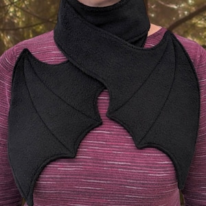 Op de afbeelding: Een zwarte fleece sjaal met bevestigde vleermuisvleugels. De sjaal wordt om de nek gedragen en de vleugels worden over de schouders gelegd.