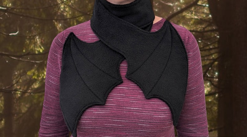Op de afbeelding: Een zwarte fleece sjaal met bevestigde vleermuisvleugels. De sjaal wordt om de nek gedragen en de vleugels worden over de schouders gelegd.