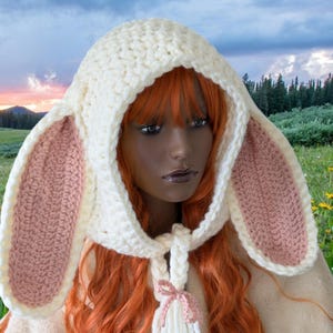 May include: A white crocheted bunny hat with pink ears. The hat has a hood and a long scarf with a tassel. The hat is worn by a person with long red hair.