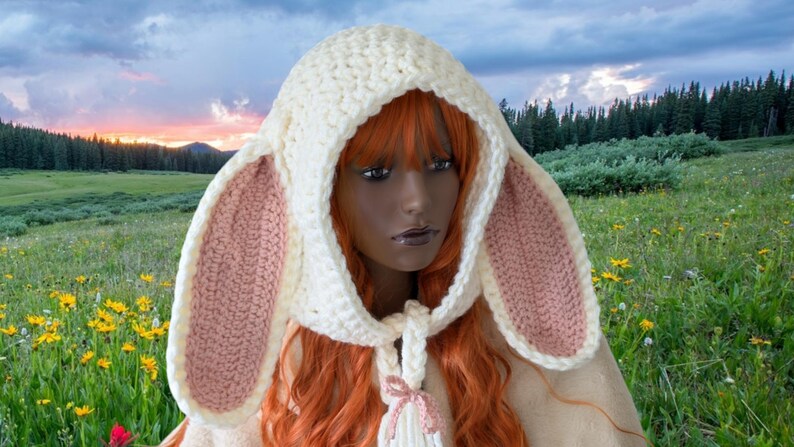 May include: A white crocheted bunny hat with pink ears. The hat has a hood and a long scarf with a tassel. The hat is worn by a person with long red hair.