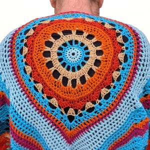 May include: A colorful crochet sweater with a large circular design on the back. The sweater features a vibrant palette of blue, orange, pink, and beige. The open weave design is visible, and the sweater appears to be handmade.