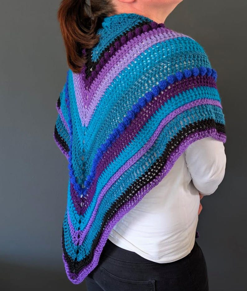 May include: A handmade crochet shawl in shades of teal, purple, and black. The shawl features a V-neck design and is adorned with textured bobbles along the edge. The color scheme creates a striking visual appeal.