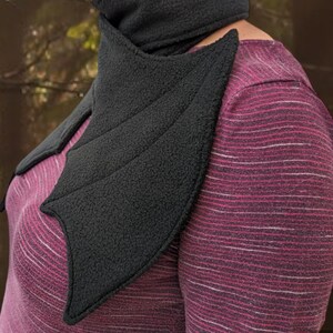 Op de afbeelding: Een zwarte fleece nekwarmer in de vorm van vleermuisvleugels. De warmer wordt gedragen over een roze en wit gestreept shirt.