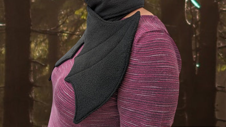 Op de afbeelding: Een zwarte fleece nekwarmer in de vorm van vleermuisvleugels. De warmer wordt gedragen over een roze en wit gestreept shirt.