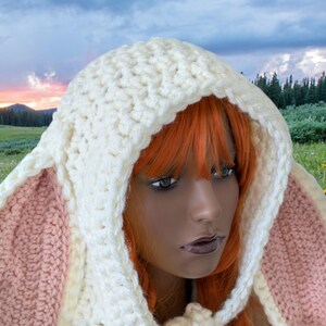 May include: A white crocheted hooded scarf with pink bunny ears. The scarf is worn by a person with long, bright orange hair. The background is a field of green grass and yellow flowers.