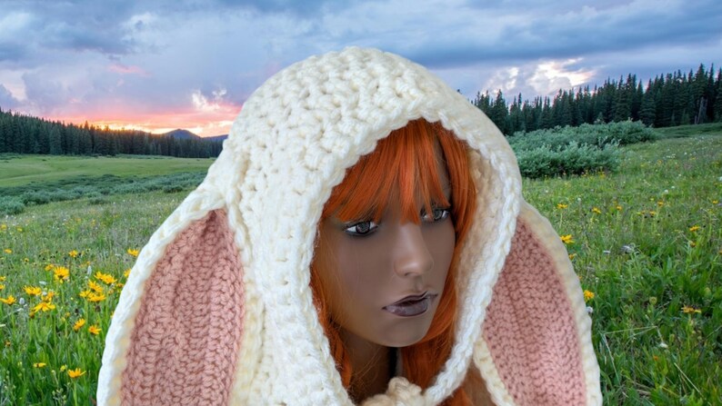May include: A white crocheted hooded scarf with pink bunny ears. The scarf is worn by a person with long, bright orange hair. The background is a field of green grass and yellow flowers.