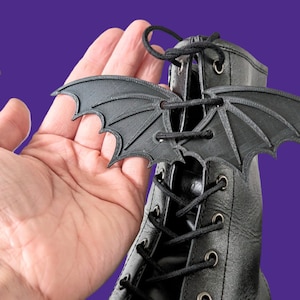 May include: Black bat-shaped shoe lace charms. The charms are made of a 3D printed material and have a detailed design. The charms are shown on a black boot with black laces.