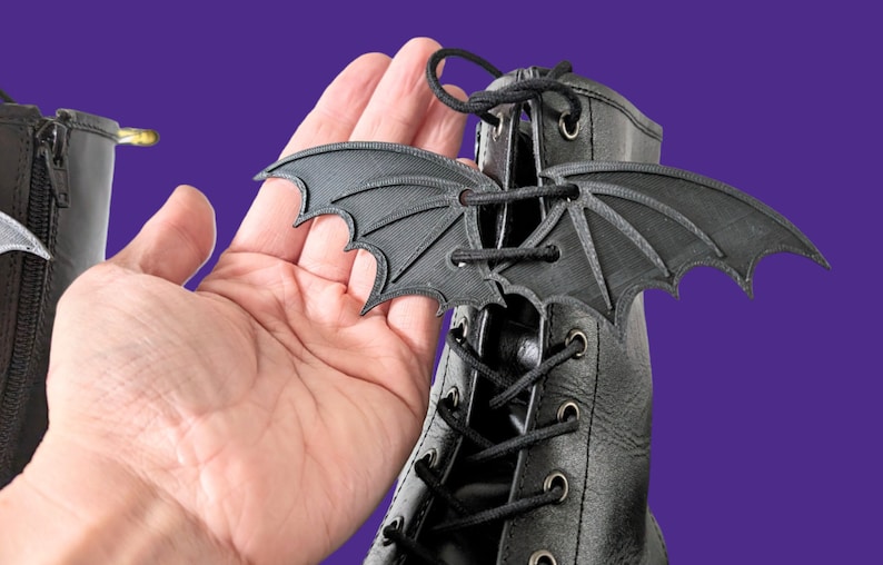 May include: Black bat-shaped shoe lace charms. The charms are made of a 3D printed material and have a detailed design. The charms are shown on a black boot with black laces.