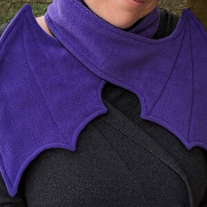 May include: A purple fleece scarf with bat wings. The scarf is worn around the neck and the wings drape over the shoulders.