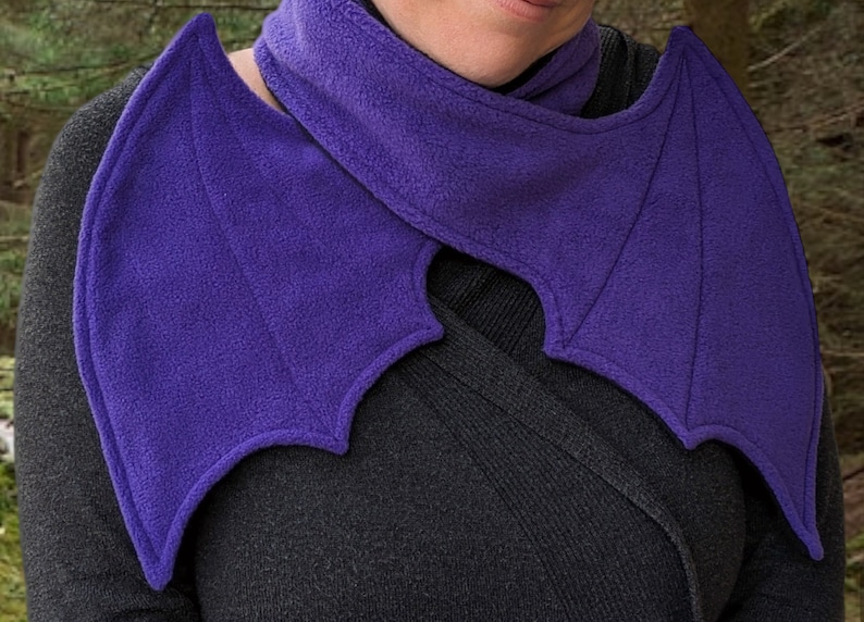 May include: A purple fleece scarf with bat wings. The scarf is worn around the neck and the wings drape over the shoulders.