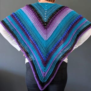 May include: A handmade, crocheted shawl in shades of teal, purple, and black. The shawl features a V-neck design with alternating stripes and textured bobble stitches. The color palette creates a striking visual effect.