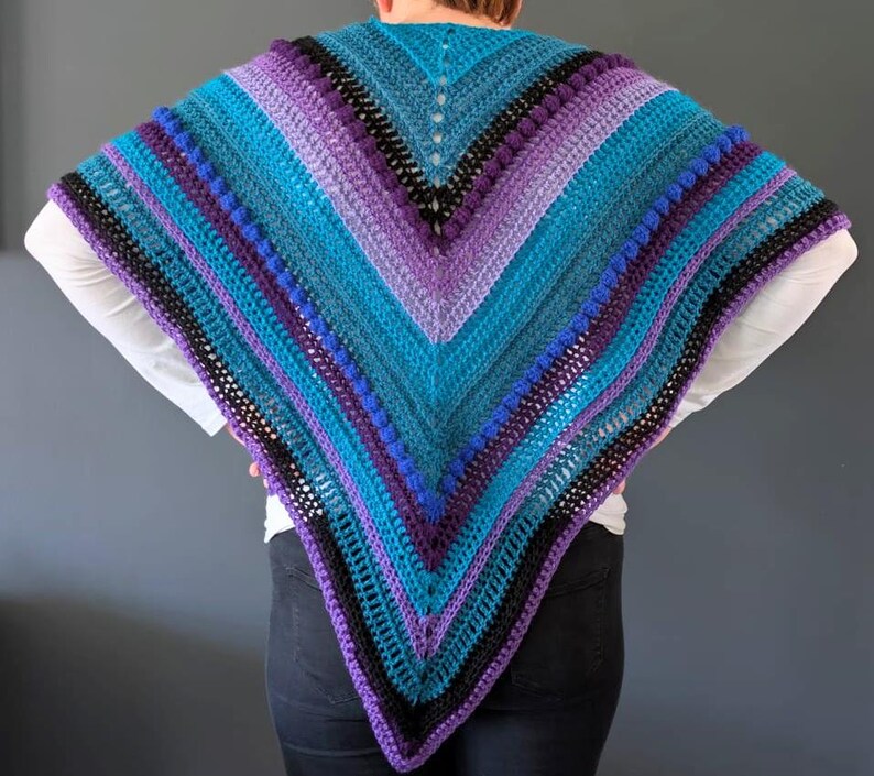 May include: A handmade, crocheted shawl in shades of teal, purple, and black. The shawl features a V-neck design with alternating stripes and textured bobble stitches. The color palette creates a striking visual effect.