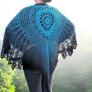 May include: A blue and black crocheted shawl with a circular design in the center. The shawl has a gradient effect, transitioning from a bright blue to a darker black. The shawl is worn over a black top and gray pants.
