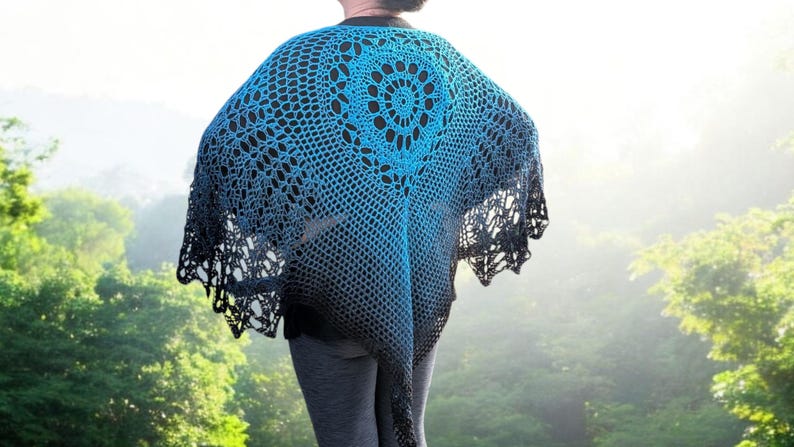 May include: A blue and black crocheted shawl with a circular design in the center. The shawl has a gradient effect, transitioning from a bright blue to a darker black. The shawl is worn over a black top and gray pants.