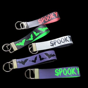 May include: Five Halloween-themed wrist keychains in various colors. Each keychain features the word "SPOOKY" or bat silhouettes. Colors include red, black, white, purple, and green. The keychains have a silver metal ring.