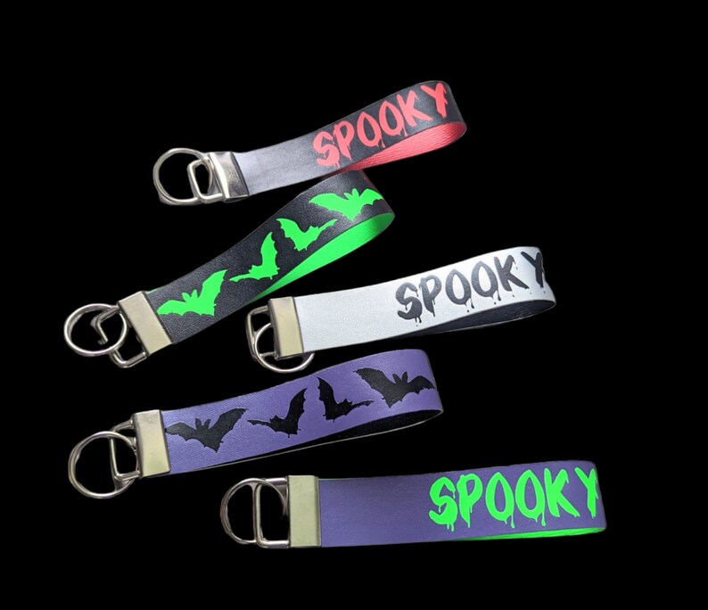 May include: Five Halloween-themed wrist keychains in various colors. Each keychain features the word "SPOOKY" or bat silhouettes. Colors include red, black, white, purple, and green. The keychains have a silver metal ring.