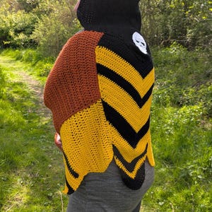 May include: A handmade crochet shawl in shades of brown, yellow, and black. The shawl features a skull applique and a chevron pattern. The person is wearing a black hood. The background is a natural outdoor setting with a path.