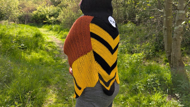 May include: A handmade crochet shawl in shades of brown, yellow, and black. The shawl features a skull applique and a chevron pattern. The person is wearing a black hood. The background is a natural outdoor setting with a path.
