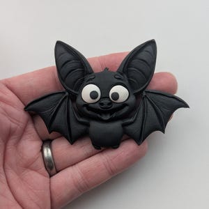 May include: A black bat figurine with large, expressive eyes and a smiling face. The bat has detailed wings and ears, and is made of a solid, matte material. The figurine is a Halloween or gothic-themed decorative item.