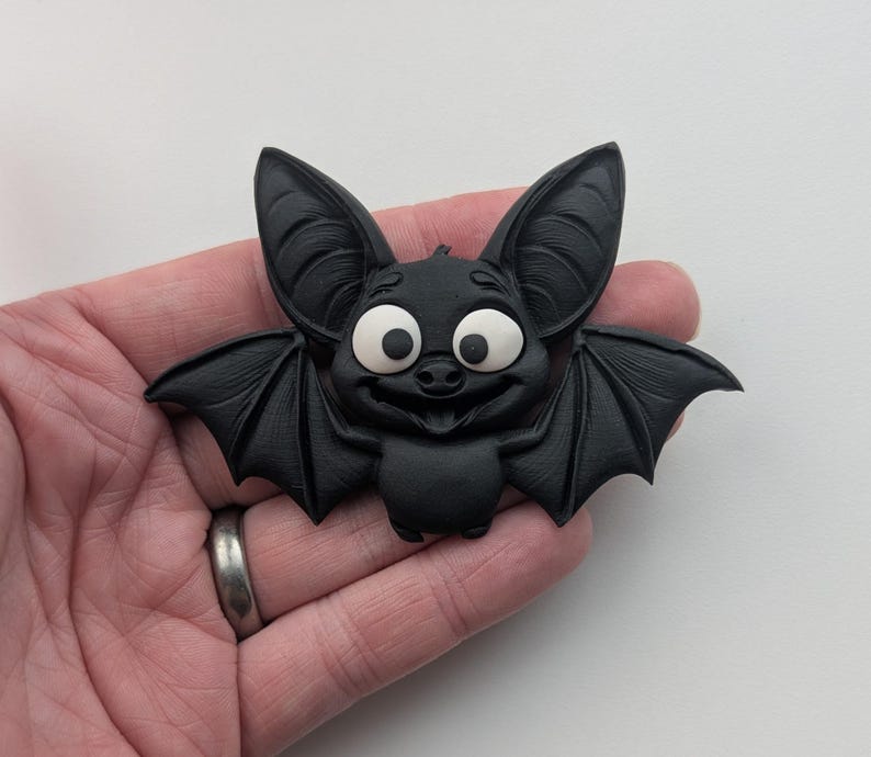 May include: A black bat figurine with large, expressive eyes and a smiling face. The bat has detailed wings and ears, and is made of a solid, matte material. The figurine is a Halloween or gothic-themed decorative item.