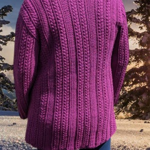 May include: A vibrant purple, long-sleeved sweater with a textured, vertical cable knit design. The sweater has a high neckline and a slightly flared hem. The person is wearing blue jeans. The background shows a winter scene.