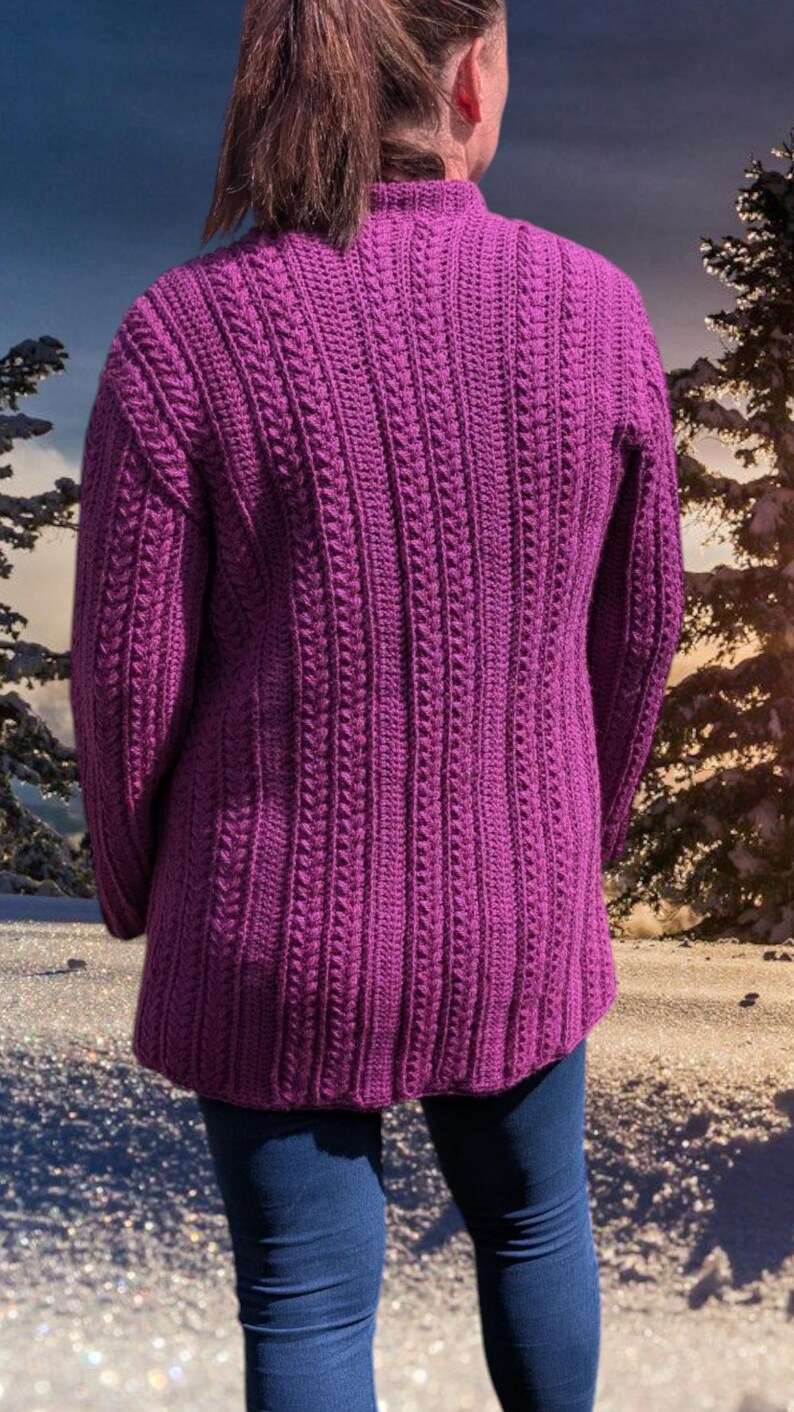 May include: A vibrant purple, long-sleeved sweater with a textured, vertical cable knit design. The sweater has a high neckline and a slightly flared hem. The person is wearing blue jeans. The background shows a winter scene.