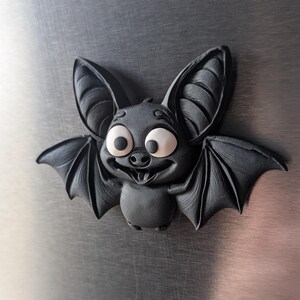 May include: A dark gray, cartoon-style bat with large, expressive eyes and a smiling mouth. The bat has large ears and outstretched wings, giving it a friendly appearance. The bat is a decorative item.