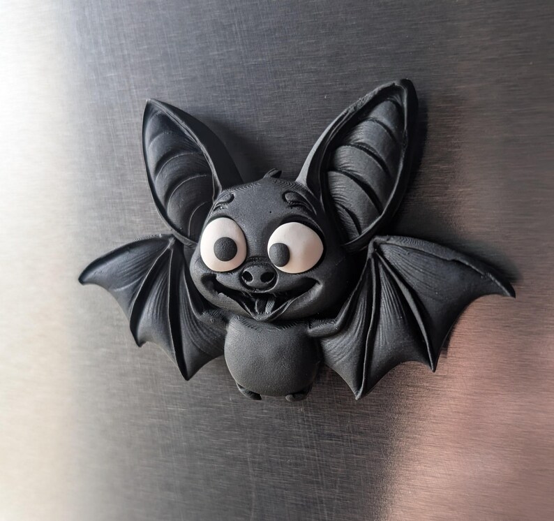 May include: A dark gray, cartoon-style bat with large, expressive eyes and a smiling mouth. The bat has large ears and outstretched wings, giving it a friendly appearance. The bat is a decorative item.