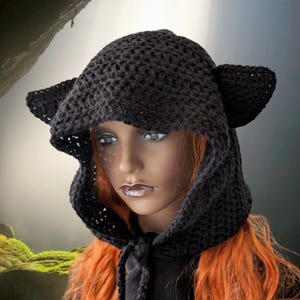 Puede incluir: Un gorro de punto negro con capucha y orejas de gato. El gorro est&aacute; hecho con un delicado patr&oacute;n de punto abierto y tiene un lazo en el cuello.