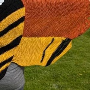 May include: A close-up of a knitted shawl with a bee wing design. The shawl features a combination of black, yellow, and orange colors. The design includes black stripes on yellow sections, and a solid orange upper portion.